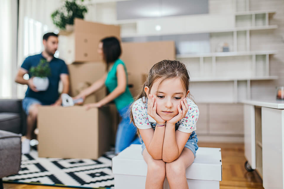 Sad girl sitting on the moving box, parents behind her 